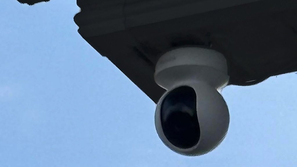 White pan-and-tilt CCTV camera fitted under a dark soffit, blue sky behind